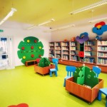 Sala działu przy wejściu, wieszak przyścienny w kształcie drzewa, przyścienne regały z książkami i pojemniki na książki na kółkach, przy których stoją dziecięce krzesełka