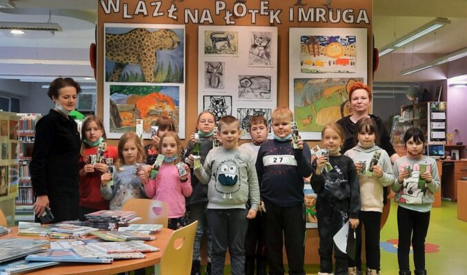 11 dzieci wraz z nauczycielką i bibliotekarką na zdjęciu grupowym przed ścianą wystawową z napisem "Wlazł na płotek i mruga, dzieci trzymają zakładki do książek