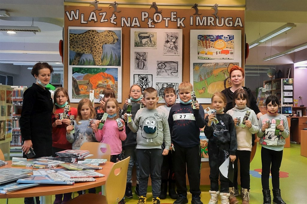 11 dzieci wraz z nauczycielką i bibliotekarką na zdjęciu grupowym przed ścianą wystawową z napisem "Wlazł na płotek i mruga, dzieci trzymają zakładki do książek
