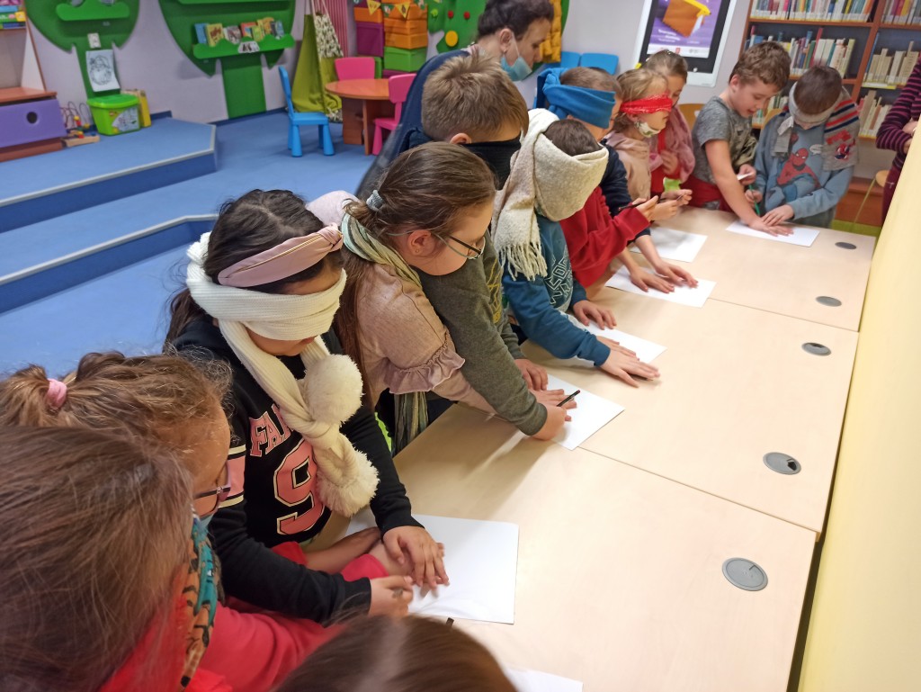 Dzieci w parach wykonują zadanie: dziecko z zasłoniętymi szalikiem oczyma obrysowuje na kartce rękę innego dziecka