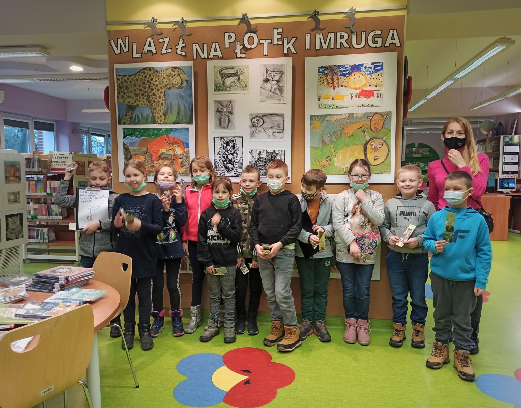 11 dzieci wraz z nauczycielką na zdjęciu grupowym przed ścianą wystawową z napisem "Wlazł na płotek i mruga, dzieci trzymają zakładki do książek