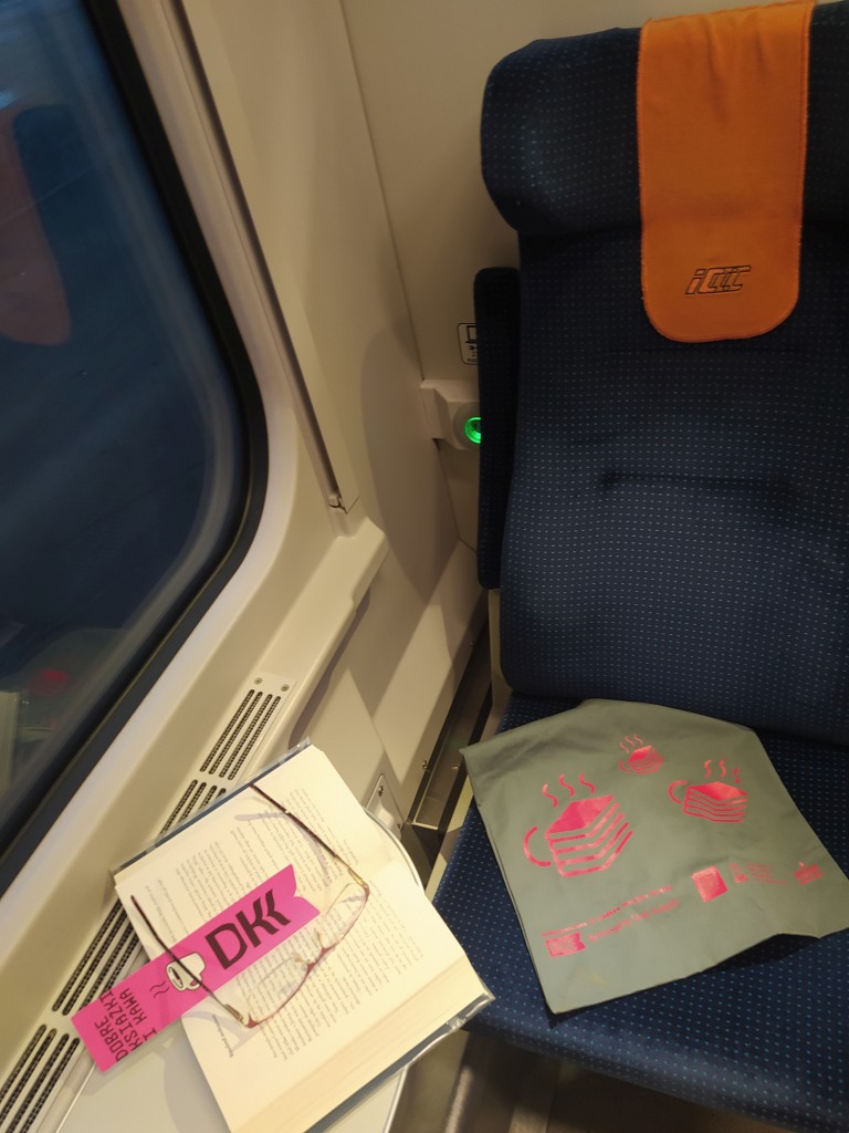 Na fotelu w pociągu ICCC leży torba firmowa DKK, obok na stoliku przyokiennym leży otwarta książka z umieszczoną na niej zakładką DKK i okularami
