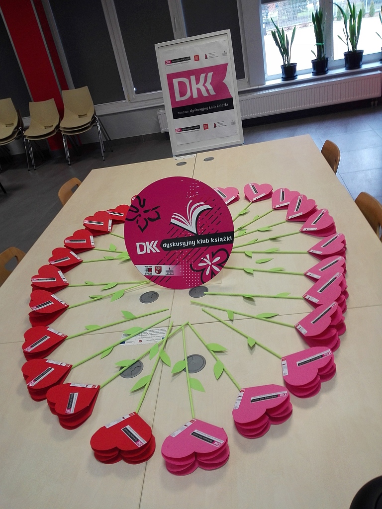 Na stole okrągła plansza z reklamą DKK, dokoła niego papierowe kwiatki z kielichami w kształcie serc i z naklejkami DKK, w tle potykacz z logiem DKK