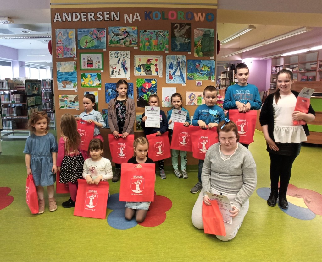 Zdjęcie grupowe laureatów na tle ściany wystawowej o tytule "Andersen na kolorowo", 12 dzieci trzyba przed sobą torby z herbem Morąga