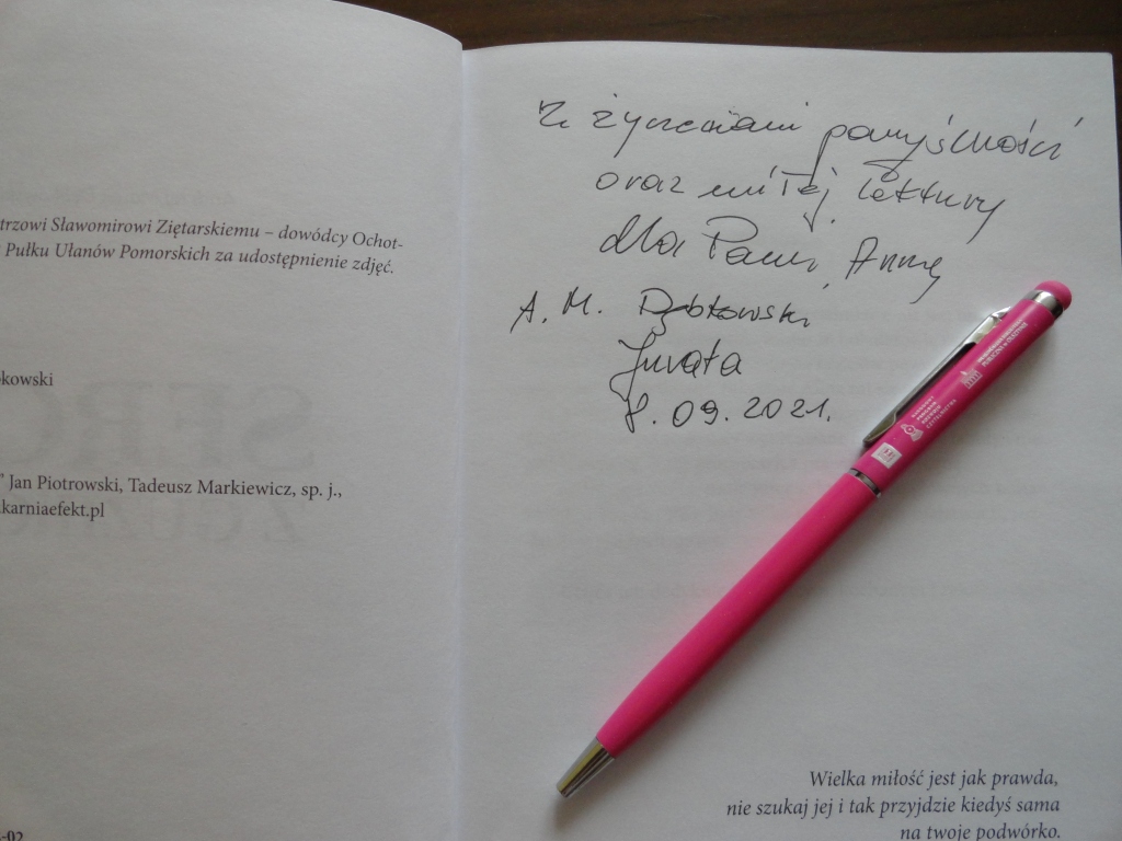 Otwarta ksiązka, na której umieszczono dedykację "Z życzeniami pomyślności oraz miłej lektury dla Pani Anny, A. M. Dąbkowski, Jurata 8.09.2021"; na książce leży różowy długopis oklejony logami związanymi z DKK