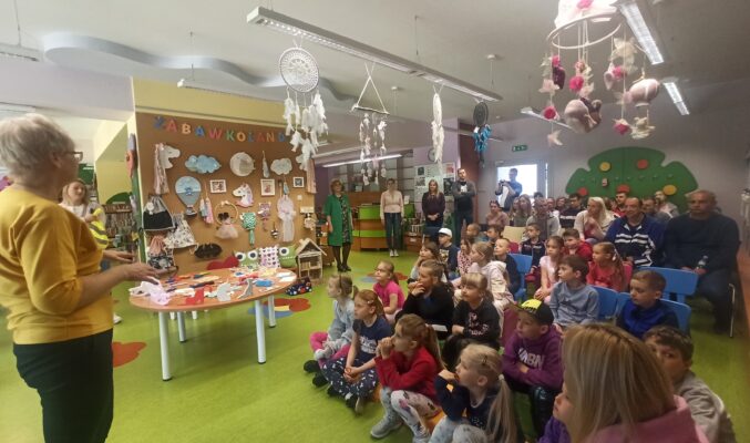 Dzieci i dorośli siedzący na krzesłach i pufach słuchają wypowiedzi dyrektorki biblioteki, w tle ściana wystawowa z wywieszonymi zabawkami i napisem "Zabawkolandia", zabawki również rozłożone na okrągłym stole i zwisają z lamp