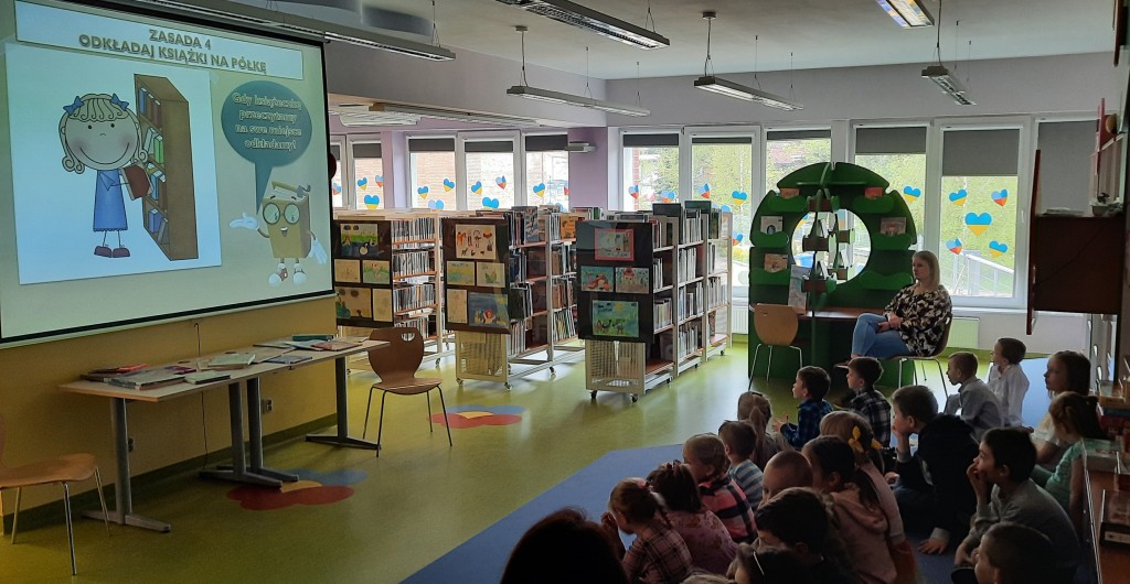 Dzieci w przyciemnionej sali bibliotecznej oglądają na ekranie zasady obchodzenia się z książką