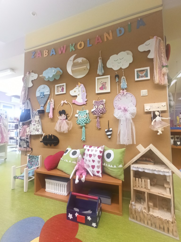 Ściana wystawowa z napisem "Zabawkolandia", na śnianie i leżace na ziemi m.in. poduszki, drewniany domek dla lalek, krzesełko, łapacze snów, torby, ramki z obrazkami