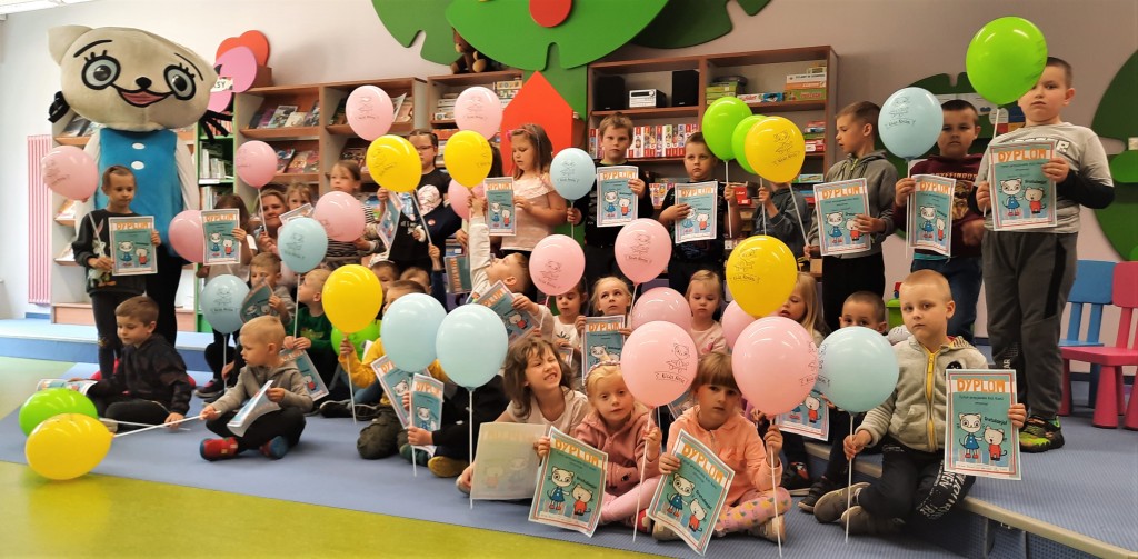 Dzieci siedzą lub stoją i trzymają w rękach kolorowe baloniki i dyplomy z Kicią Kocią, z tyłu po lewej stoi osoba w przebraniu Kici Koci, za nimi regały z książkami, czasopismami, grami i puzzlami