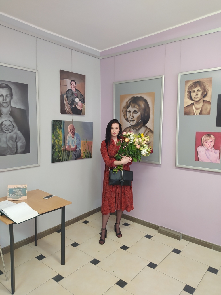 Główna autorka wystawy stoi z bukietem kwiatów w rogu pomieszczenia pomiędzy obrazami, obok stoi kwadratowy stolik z księgą pamiątkową