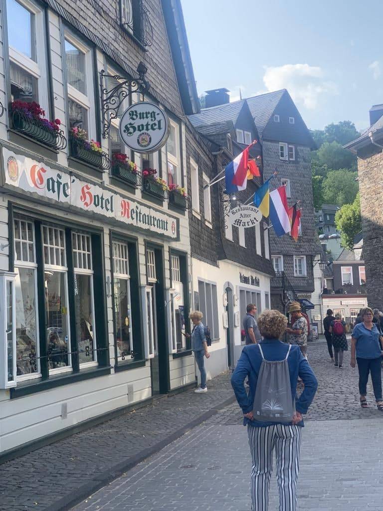 Ulica z zabytkowymi alpejskimi domami, jeden o nazwie "Burg-hotel", starsza pani stoi na ulicy odwócona tyłem, na plecach nosi firmową torbę DKK