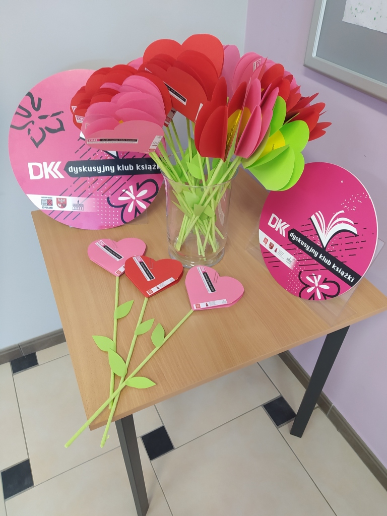 Na kwadratowym stoliku leżą 3 papierowe kwiaty w kształcie serca, wazon z dużą ilością tych samych kwiatów oraz 2 okrągłe plansze z logiem DKK