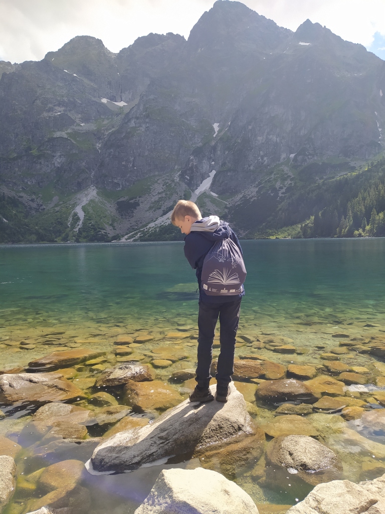 Chłopiec z firmowym plecakiem DKK stoi na kamieniu na brzegu górskiego jeziora
