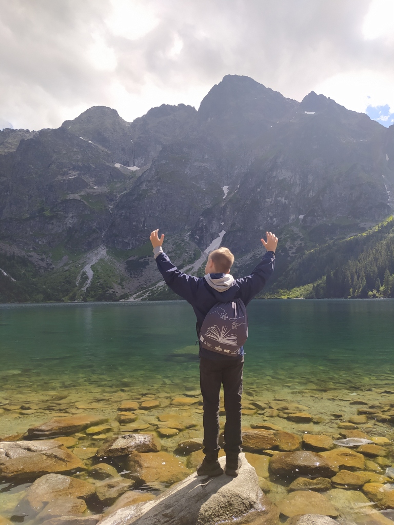 Chłopiec z firmowym plecakiem DKK stoi na kamieniu na brzegu górskiego jeziora i wnosi do góry ręce