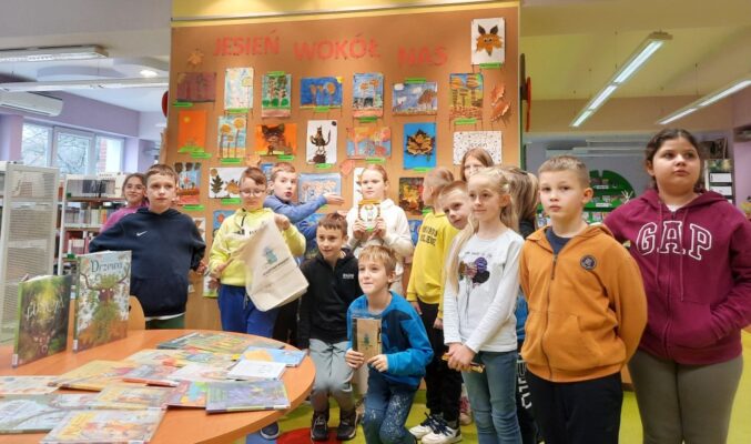 Dzieci na tle ściany wystawowej z pracami o tematyce "Jesień wokół nas", niektóre dzieci trzymają książki, przed dziećmi stół z rozłożonymi książkami