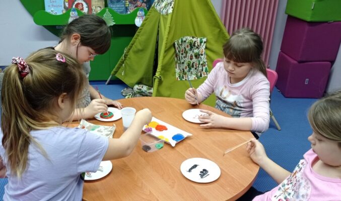 4 dziewczynki siedzą dookoła okrągłego stołu i malują farbami na papierowych talerzykach