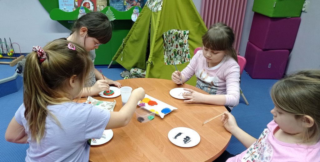 4 dziewczynki siedzą dookoła okrągłego stołu i malują farbami na papierowych talerzykach
