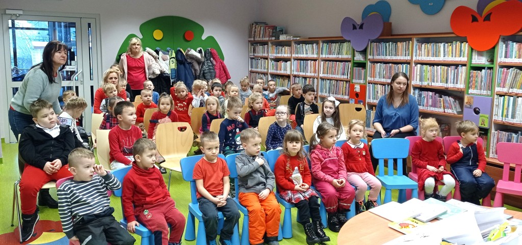 Ok. 30 dzieci siedzi na krzesłach, po prawej przy ścianie ciąg regałów z książkami