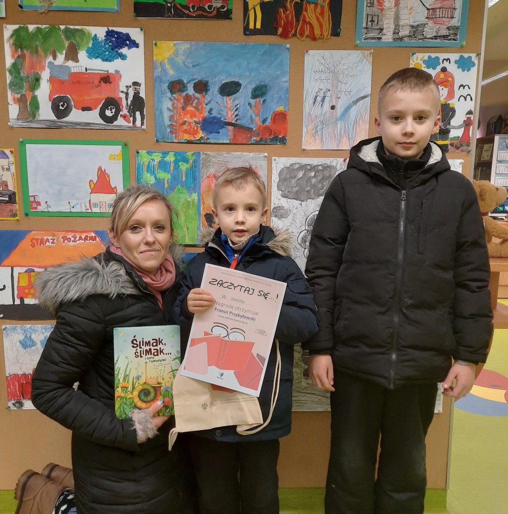 Laureat z stycznia 2023 trzyma w rękach torbę i dyplom, po prawej stoi jego brat, po lewej klęczy mama i trzyma książkę "Ślimak, ślimak…"