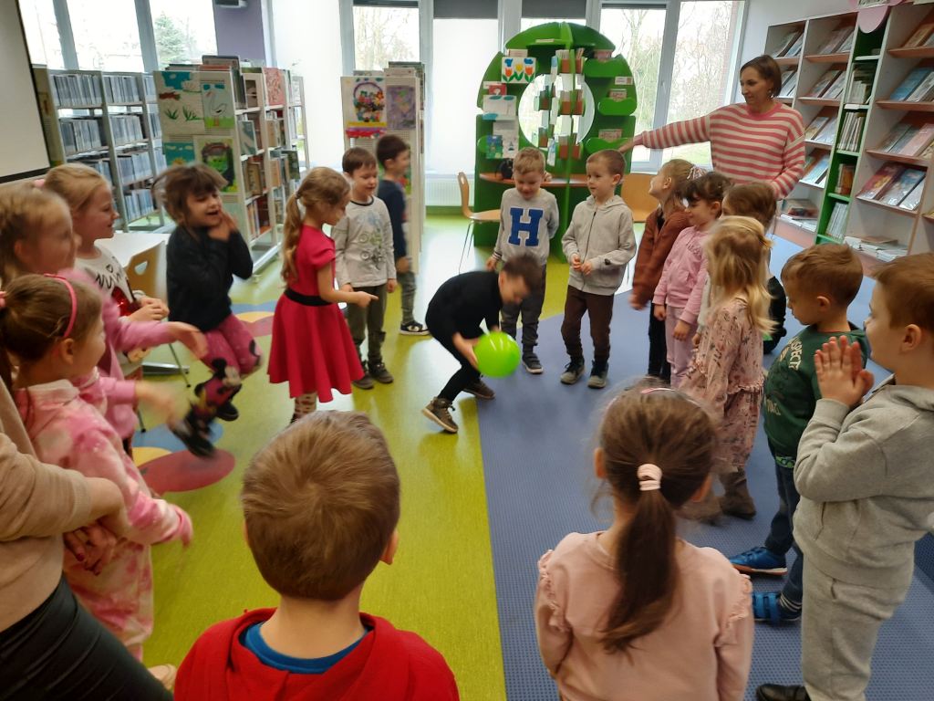 Dzieci stoją w kółku, część dzieci podskakuje, chłopiec pośrodku łapie zielony balon, obok stoi opiekunka i wskazuje ręką na innego chłopca