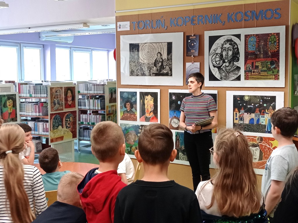 Bibliotekarka trzymając książkę opowiada o wystawie, za nią ściana wystawowa zatytułowana "Toruń, Kopernik, Kosmos", z przodu sylwetki dzieci przysłuchujących się