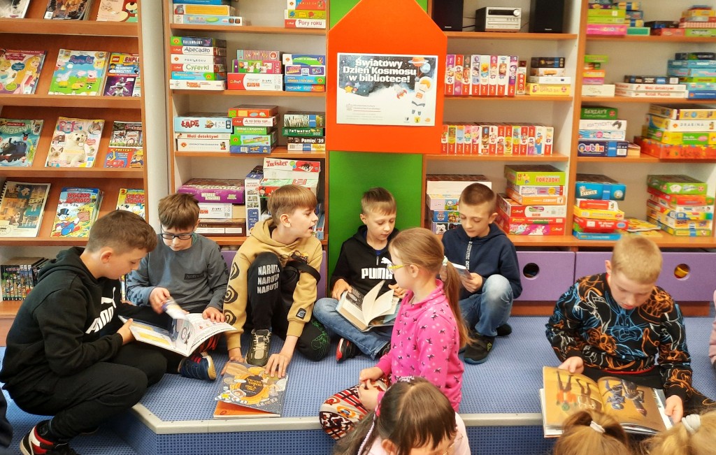 Dzieci siedzą po turecku lub kucają i w grupkach przeglądają książki, za nimi regały z czasopismami, grami i puzzlami