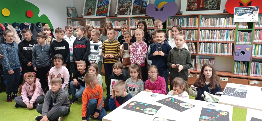Ok. 30 dzieci pozują do zdjęcia na tle regałów z książkami, w pobliżu stoją stoły, na których ułożono prace plastyczne wykonane na zajęciach