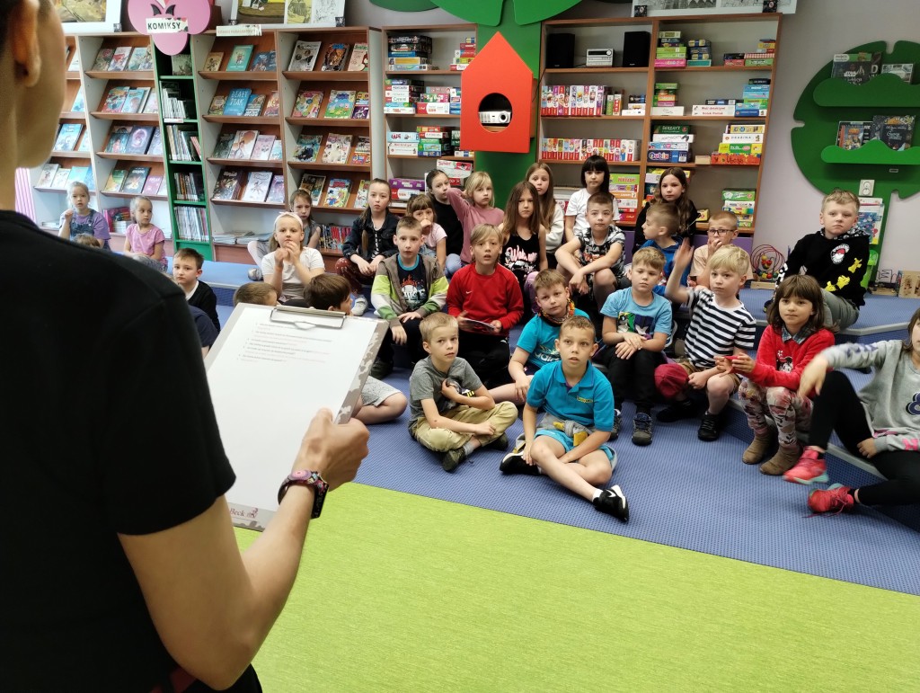 Bibliotekarka czyta dzieciom z kartki zagadki, dzieci siedzą na podłodze lub stopniach i słuchają, jeden chłopiec podnosi do góry rękę