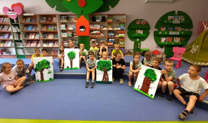 Dzieci siedzą w 4 grupach na stopniach, każda grupa trzyma dużą kartkę z narysowanym i wyklejonym drzewem, w tle regały z grami, puzzlami, czasopismami i komiksami