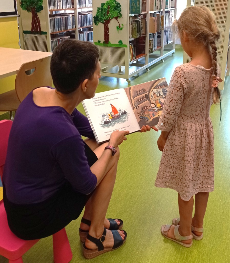 Bibliotekarka siedzi na krzesełku i pokazuje stojącej obok dziewczynce ilustracje z książki