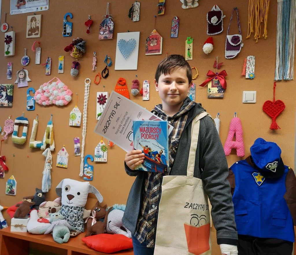 Laureat z maja 2023 stoi na tle ściany wystawowej z zabawkami i gadżetami i trzyma w ręce dyplom i książkę "Mazurscy w podróży", a na ramieniu trzyma bawełnianą torbę z logo konkursu