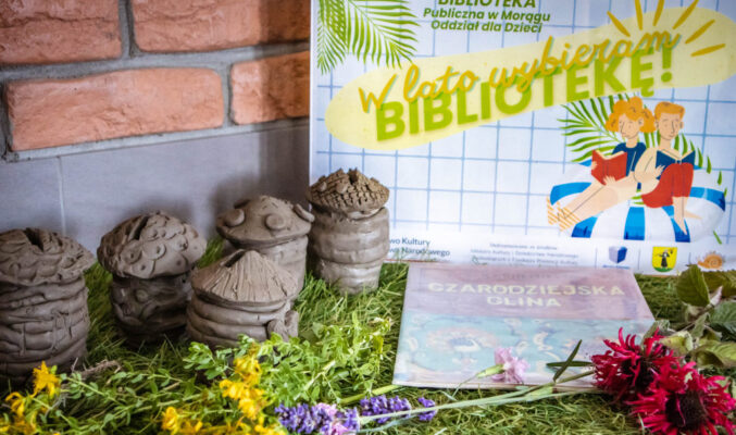 Na wykładzinie przypominającej trawę przy ceglanej ścianie leżą: plakat warsztatów "W lato wybieram BIBLIOTEKĘ!", kolorowe kwiaty, książka "Czarodziejska glina" i 5 skarbonek wykonanych z gliny