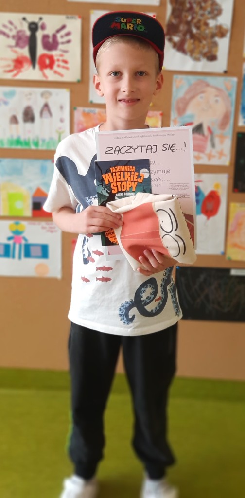 Laureat z czerwcaa 2023 stoi na tle ściany wystawowej z rysunkami dzieci i trzyma w rękach dyplom, książkę "Tajemnica Wielkiej Stopy" i złożoną bawełnianą torbę z logo konkursu
