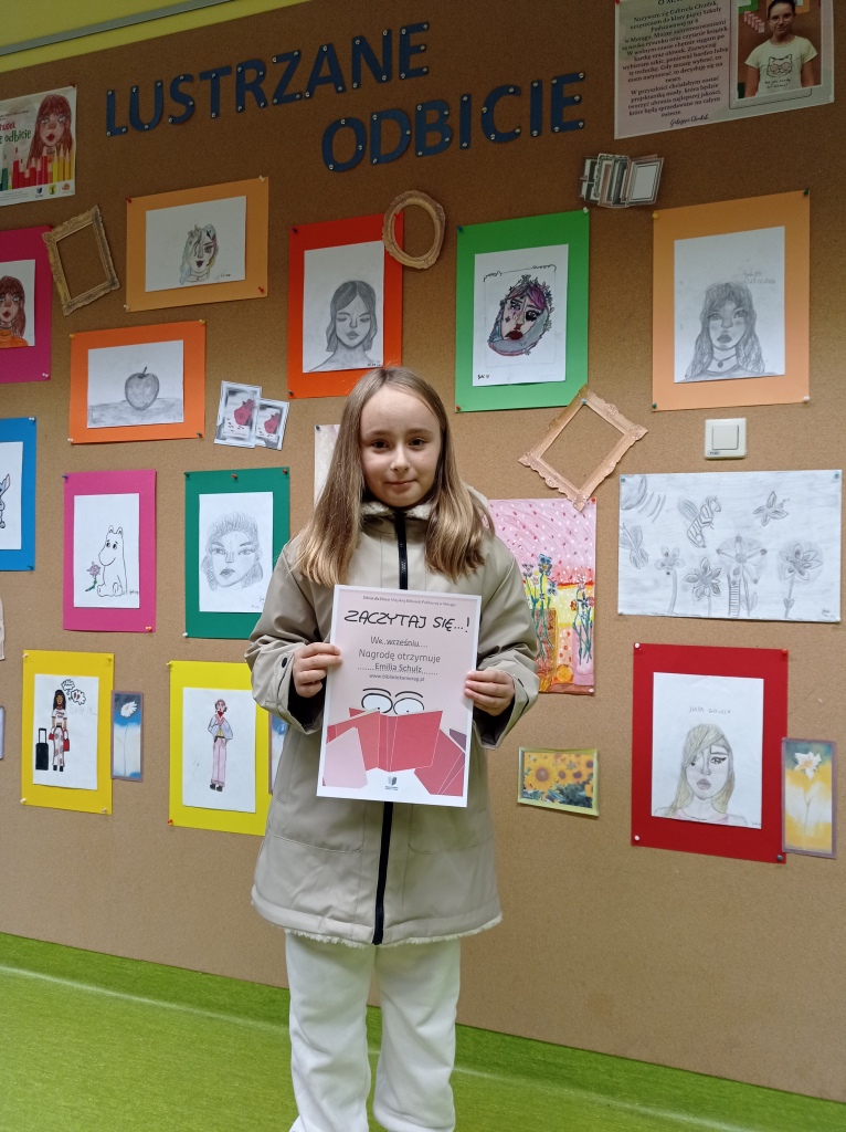 Laureatka z września 2023 stoi na tle ściany wystawowej z pracami dzieci ściany i trzyma w rękach dyplom