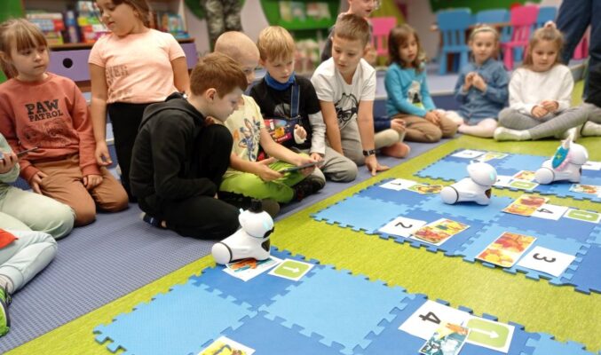 Roboty Photony poruszają się po planszach, obok dzieci siedzą na podłodze i obserwują, niektóre dzieci trzymają tableta